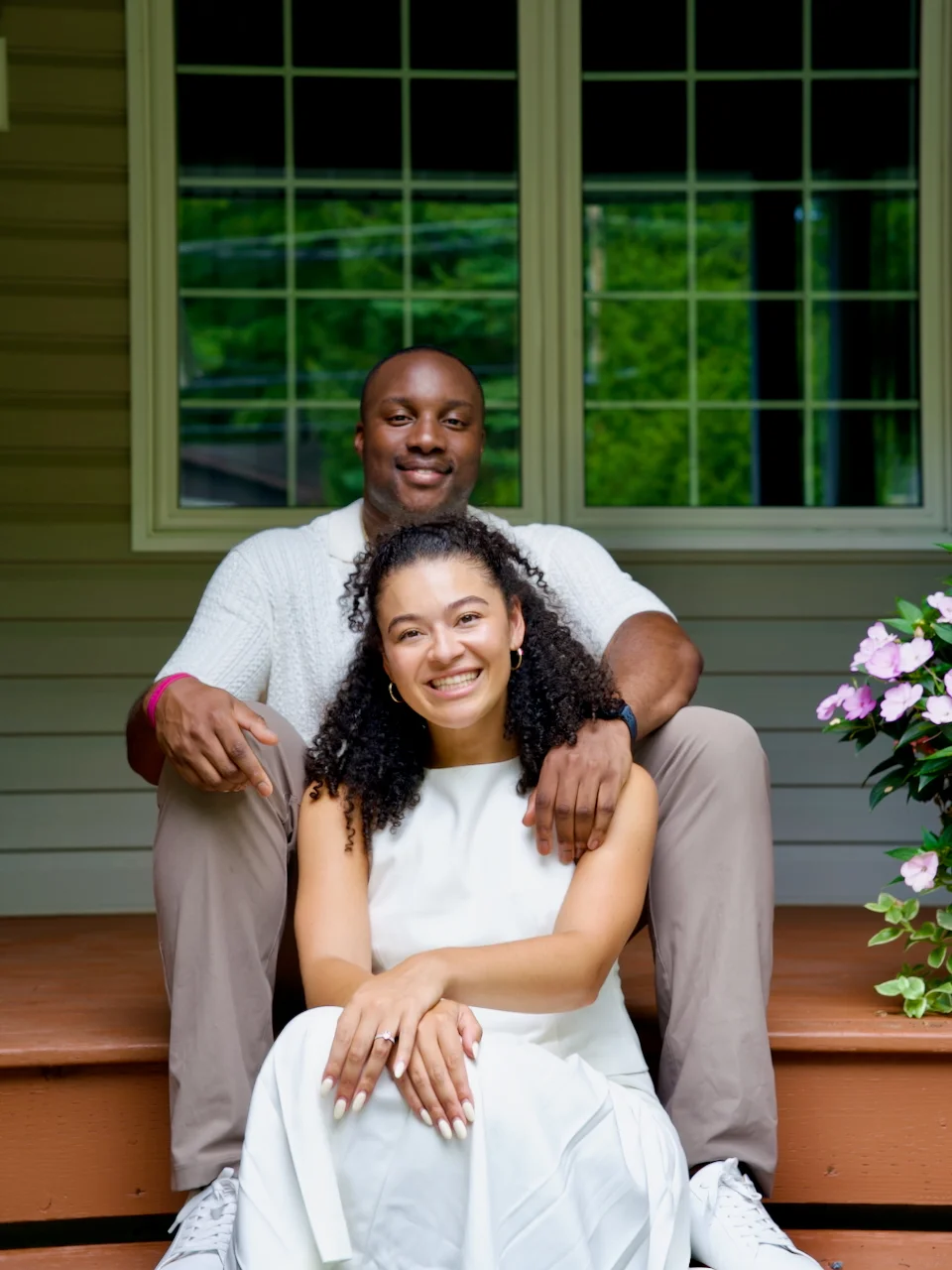 An engaged couple embracing in a backyard garden, the man standing behind the woman with his arms around her as both smile at the camera.